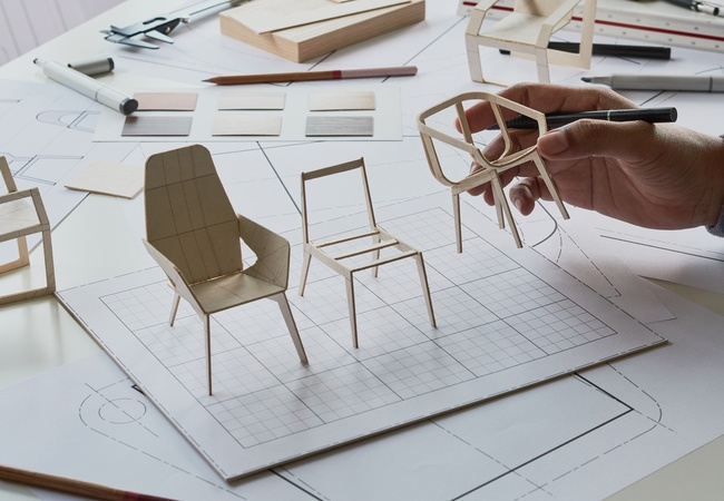 A designer holding one of three chair prototypes on their desk, each with different shapes, sizes, and styles.