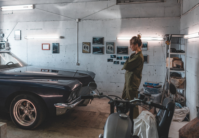 A person in overalls standing in their garage workshop and admiring the black muscle car parked in front of them.